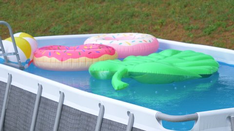 Small backyard pool is left full of colorful floaties during a late summer rainstorm. Strong stormy winds move colorful inflatable toys around the empty garden pool. Pool is abandoned during a storm.
