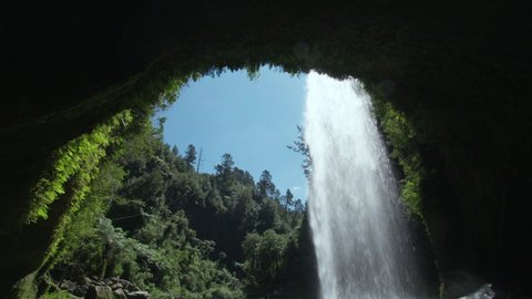 Inside cave view behind powerful Omanawa waterfall in New Zealand on sunny day
