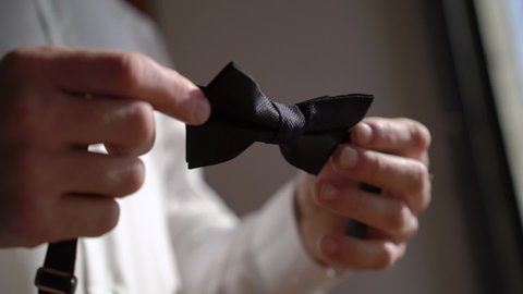 A young man is holding a black bow tie. Groom dresses adjusting for an event or wedding.