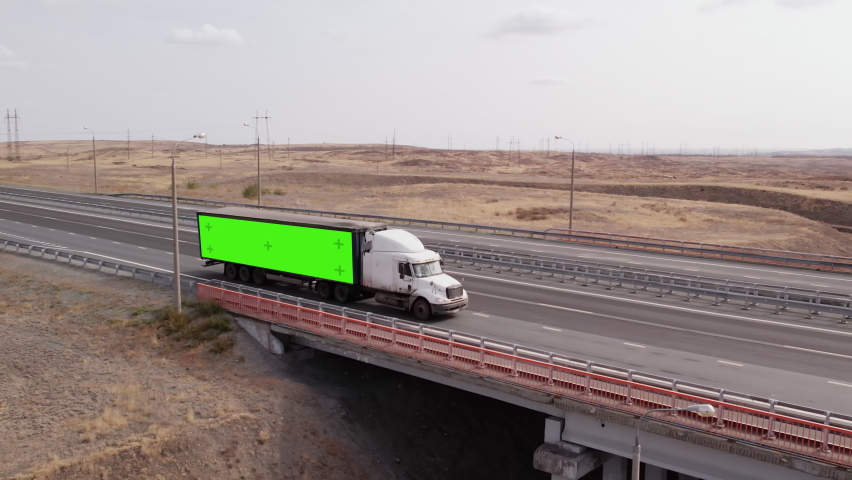 Truck with semi-trailer on which green screen passes over bridge on highway, aerial view. Truck with semi-trailer which chromakey, rides freeway
