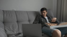 Young man is sitting on sofa eating slice of pizza from delivery cardboard box and watching video on laptop. Man is eating pizza with black dough. Modern european interior in apartment.