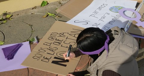 woman making a poster on the ground for feminist protest outdoors. young woman writing "We are the cry of those who are not" in Spanish on corrugated cardboard. 