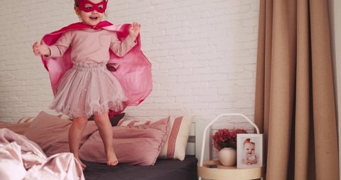 happy toddler baby girl in super hero outfit jumping on the bed on sunny day at home