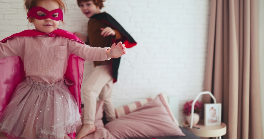 happy kids, siblings in super hero outfits having fun, jumping on the bed on sunny day at home