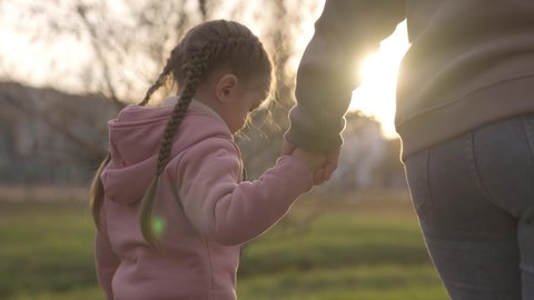 child holds her mother's hand in the rays of light in the park, little girl goes for walk with her mother to playground, concept of happy family life, kid at evening sunset with an adult is cheerful