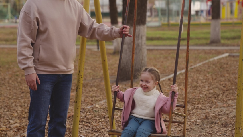 mother shakes little child on playground swing, cheerful kid flies up and down, baby laughs and smiles while playing, mother and daughter on walk in city park, happy family, childhood dream of flying Royalty-Free Stock Footage #1084743271
