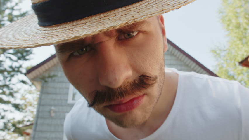 Indignant look and approaches closely into the camera of a young funny mustachioed man in straw hat in front of his house. Cinematic wide angle lens closeup face portrait with distortion