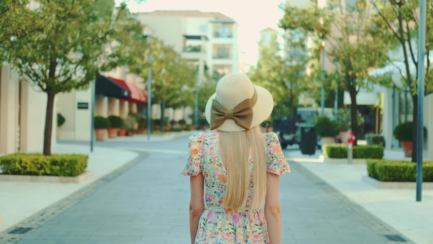 Back view of slim blonde woman in floral dress and summer hat walking down the street. Beige hat with brown ribbon bow.