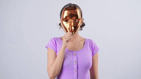 A young modern Indian Asian suspicious female hiding her face behind a mask looking at the camera isolated on white background. concept of false identity, scammer, honey trapping, or anonymity hacking