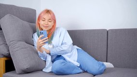 Happy young woman using smart phone with smile.Cute white girl with dyed hair browsing social media app on modern mobile phone on couch.4k stock video of Caucasian female browse internet on cellphone
