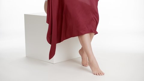 Horizontal medium shot of good-looking plus size woman in white underwear throw a piece of red silk cloth down her smooth legs sitting on white cube podium on white background | Smooth legs concept