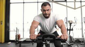 Portrait of a sportive brunette man doing cardio on a exercise bike at gym. On the background is a large window. Moving away video.