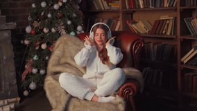 Christmas music. Young girl in headphones sits leather armchair near Christmas tree listens music and dances dressed white pajamas with hooded ears Pretty caucasian female enjoy music and have fun