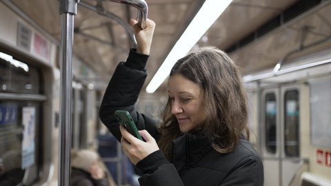 Brunette-haired woman wearing warm puffy jacket scrolls through social media using smartphone and holding on to handrail in subway wagon closeup