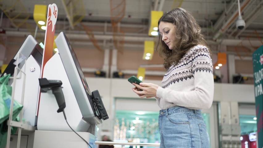 Brunette wavy-haired young woman wearing jeans and warm patterned sweater pays at self-checkout point using pay pass of smartphone Royalty-Free Stock Footage #1084770352