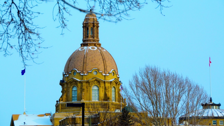 Alberta Legislature during winter twilight mid afternoon with partially snow covered roof Canadian and provincial flag at full mass trees overlooking foreground on a cold winter day prior to closures