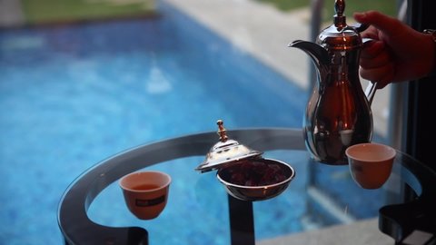 Hand with Arabic teapot pouring tea into cups. Pool in background. Gimbal, rack focus