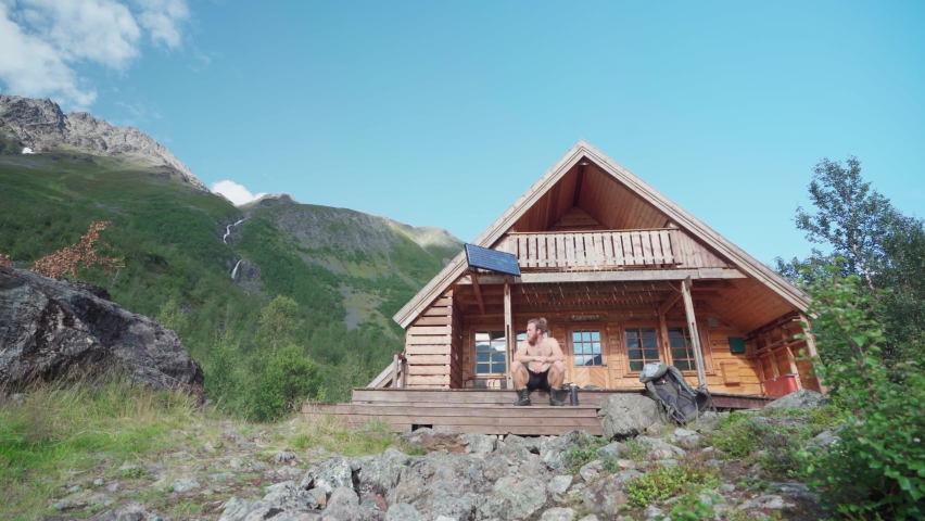 Shirtless Man Tourist Relaxing Sitting On The Front Porch Steps Of A Wooden Cabin In Lyngsdalen Valley, Norway. - Low-Angle Shot