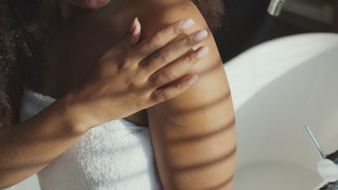 After bath skin care. Young unrecognizable black woman wrapped in towel applying moisturizing cream on her shoulder, sitting on bathtub, tracking shot, slow motion