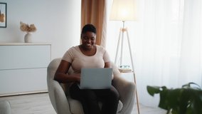 Overweight African American Woman Using Laptop Watching Movie Having Fun Sitting In Chair At Home. Happy Lady Typing And Browsing Internet On Computer Working Online Indoors. People And Technology