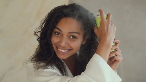 Hair Care cosmetics. Young positive african american woman spraying special conditioner on her curly care, making hairstyle at home in morning, mirror pov portrait, slow motion