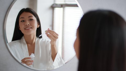 Facial Skincare. Happy Korean Lady Applying Facial Serum Using Dropper Standing In Modern Bathroom. Female Moisturizing Caring For Skin Using Natural Oils Looking At Her Reflection In Mirror Indoor