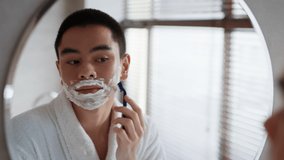 Reflection Of Asian Man Shaving Using Safety Razor Standing Near Mirror In Modern Bathroom At Home. Male Removing Facial Hair With Shaver, Having Face Covered With Foam Indoor. Selective Focus