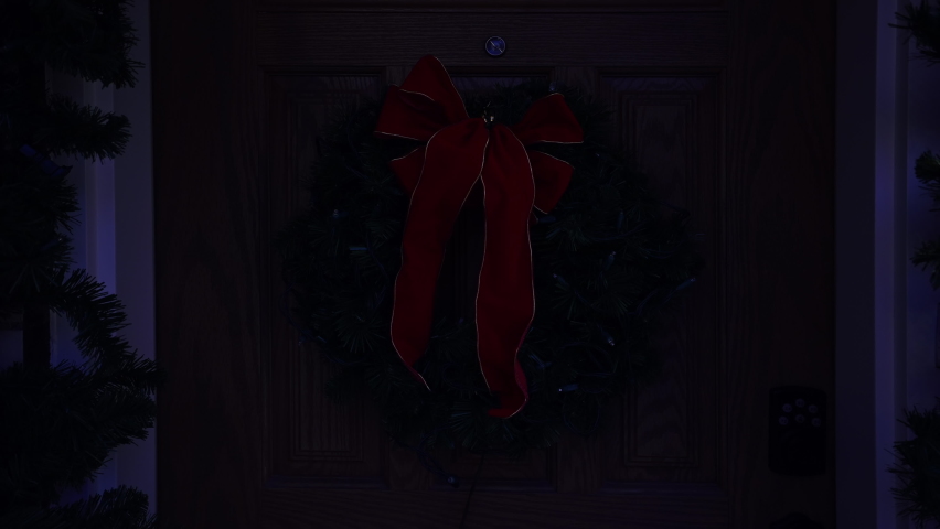 A large Christmas wreath on the front door of a typical house lights up.  	
