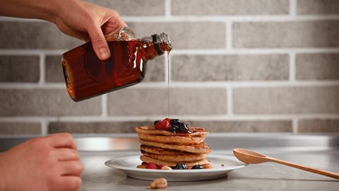 Hands of chef confectioner dripping maple syrup onto a stack with delicious homemade pancakes beautifully served with berries on a white dish. Food art. Shrovetide, Shrove Tuesday concept. Close-up.