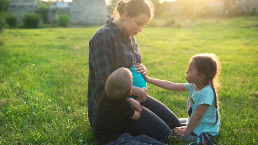 Summer, nature, happy family, pregnancy - young pregnant woman mother with two small toddler children sit on grass in park at sunset. Kids stroking mom belly kiss have fun, laugh spend time together | Shutterstock HD Video #1084789687