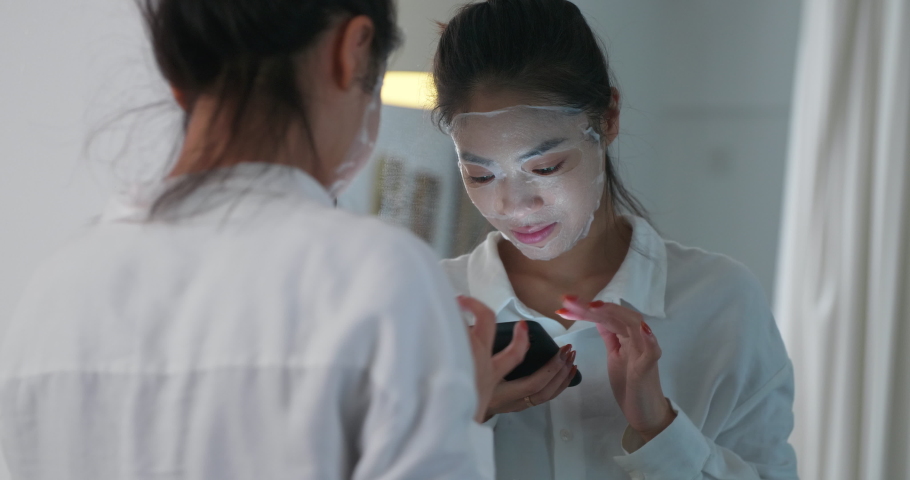 Young Asian woman wearing a facial mask looking in the mirror while looking at the phone, sending messages and browsing the web online Girl beauty skin care lifestyle at home