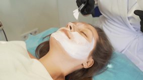 Cosmetologist applies mask to skin of woman's face for therapeutic purposes. Patient is lying on couch and thick white substance is applied to face with brush. Rejuvenation, acne treatment