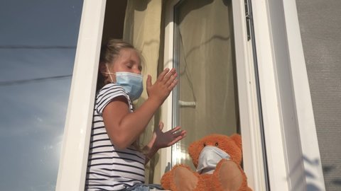 kid in medical mask sits by an open window.toddler girl applauds pandemic doctors.kid thanks medical staff fighting Covid-19.kid applauds his hands to doctors in street, looks out window wearing mask.
