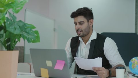 Young indian man confused by mistake in documents, looking through papers, frowning using laptop, failing urgent task, missing deadline, quits after bad work fed up with difficult job.