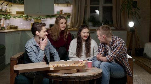 a company of four young guys and girls clink glasses cardboard cups at a home pizza party. Man with amputated arms celebrates birthday with friends in living room