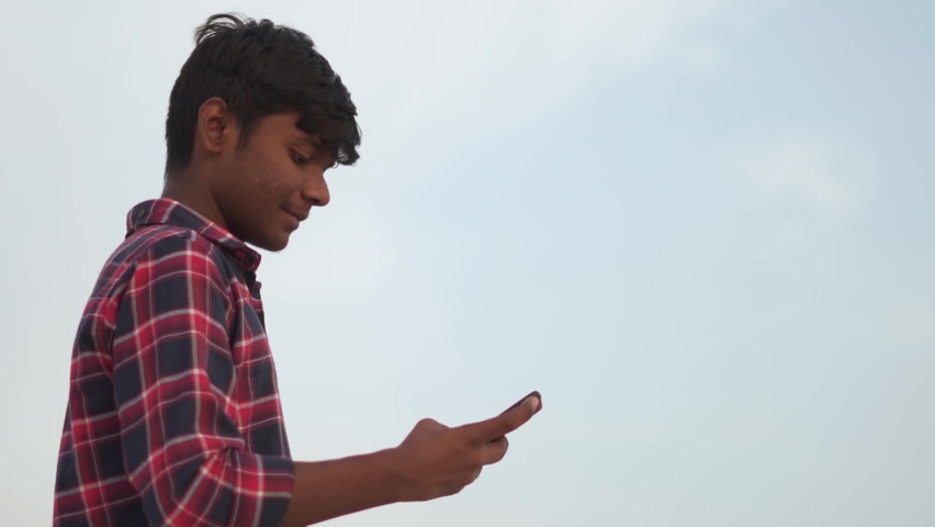 Portrait of an Indian teenager scrolling through the mobile phone in front of the sky. Happy teen swiping through the mobile phone. Social media addiction amongst the teenagers.  | Shutterstock HD Video #1084817626