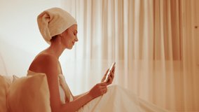 Beauty procedures at spa resort. Happy lady in white gown with towel turban talks at video call via cellphone on bed in hotel room