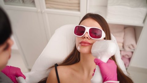 Professional cosmetologist makes a laser facial hair removal to a beautiful woman in a beauty salon. Beautician and spa concept. Body care. Slow motion. Close-up