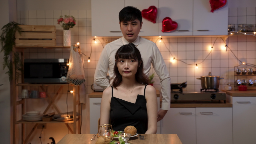 surprised asian woman smiling and smelling the red rose she gets from boyfriend on valentine’s night date at home. the man covers girlfriend’s eyes and kissing her as he gives flower