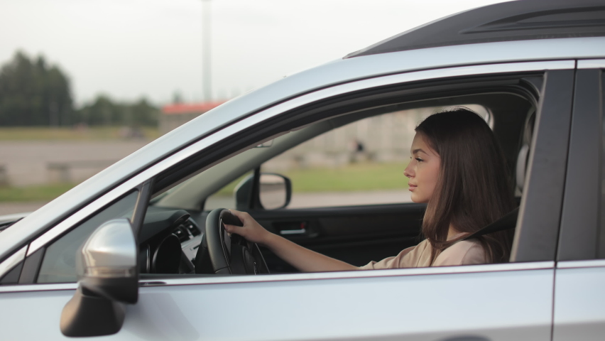 A young woman is sitting in the car and smiling. She is turning her head and looking at the camera. She is keeping her hands on the steering wheel. The camera zooms in. 4K