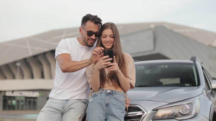 A man and a girl are standing near a car. They are smiling and watching something on their smartphone. The camera is moving around them. 4K