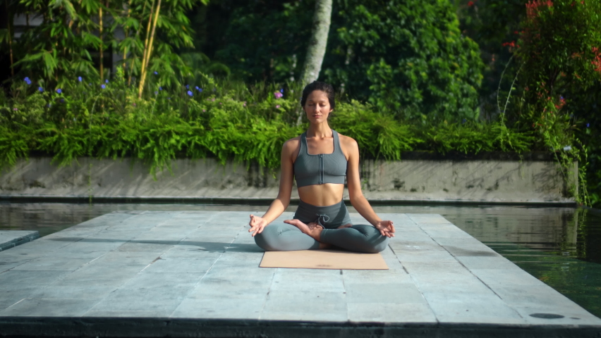 A young attractive European brunette sports girl makes yoga in nature with a green rainforest on a lit background by the pool on a mat and breathes deeply.