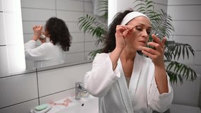 Attractive beautiful woman in a white waffle robe standing with her back to the mirror in the bathroom and making eyebrow correction, plucking eyebrows looking at herself in a small cosmetic mirror.