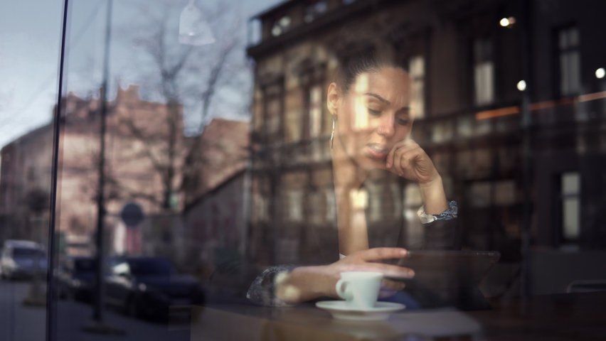 Portrait through window glass of young pensive business woman drinking coffee in a coffee shop on her work break and looking in the tablet, city reflections on window Royalty-Free Stock Footage #1084837093