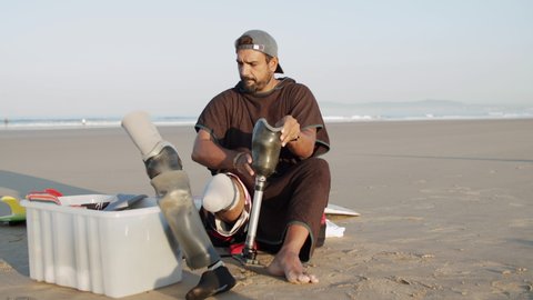 Front view of pensive surfer putting on prosthetic leg on coast. Long shot of focused bearded man in cap preparing for surf training in ocean at sunset. Disability, surfing concept