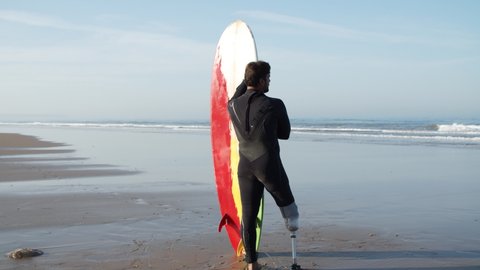 Back view of surfer with bionic leg in wetsuit looking at ocean. Long shot of sportsman with disability standing on sandy shore with paddleboard, admiring seascape. Disability, surfing concept