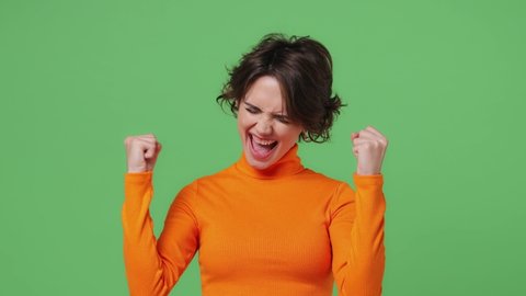 Excited jubilant overjoyed happy vivid young brunette woman 20s years old wears orange shirt doing winner gesture celebrate clenching fists say yes isolated on plain green background studio portrait