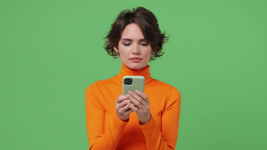 Happy young brunette woman 20s wear orange shirt hold use mobile cell phone typing say wow yes just found out great big win news doing winner gesture isolated on plain green background studio portrait Royalty-Free Stock Footage #1084838980