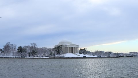 The Jefferson Memorial and Tidal Basin in Washington, D.C. as seen on a snowy winter day. Two distant people walk in front of the memorial. The sky is overcast. Geese fly by in the sky.