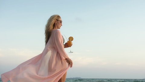 Woman walks on sunset on beach against background of sea. Girl in swimsuit and pink shirt cape drinks her orange cocktail